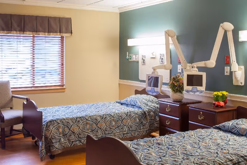 Two twin beds with patterned bedspreads in a clean, well-lit senior care room with bedside tables, monitor arms, a window with blinds, and a chair.