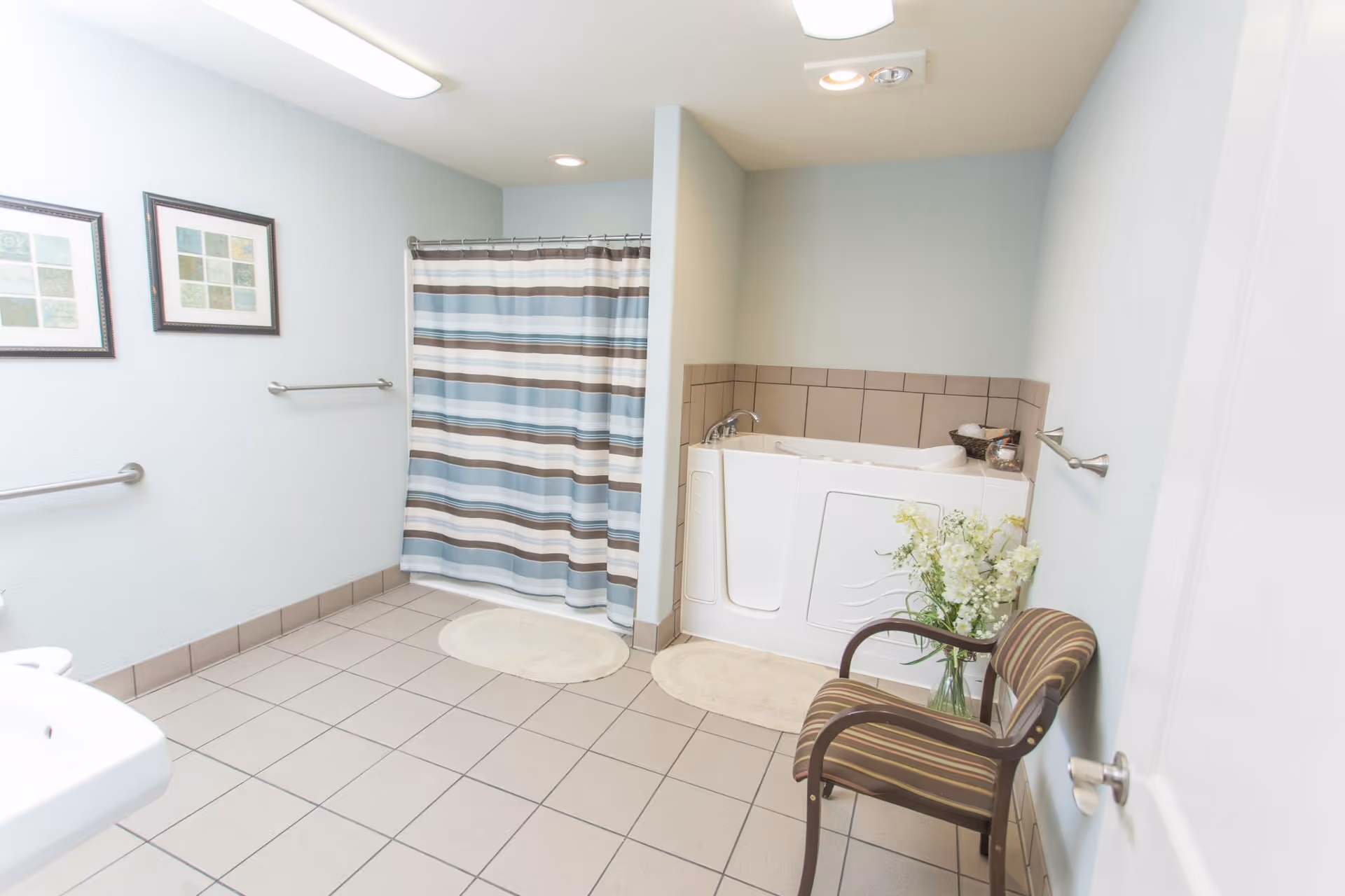 Accessible bathroom with a walk-in tub, shower with a striped curtain, tiled floor, sink, grab bars, and a striped chair with flowers.