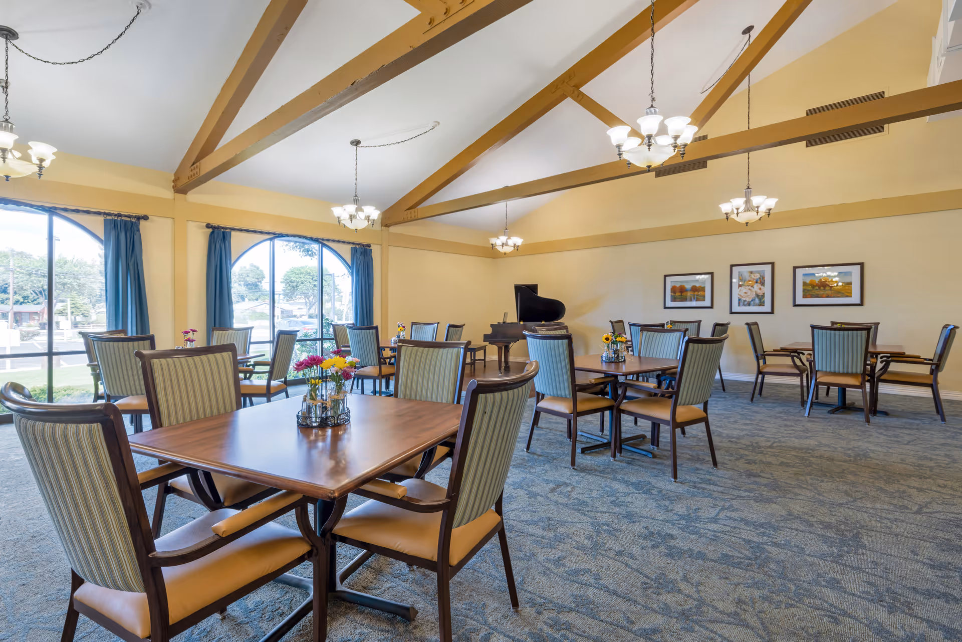 A spacious dining room with multiple wooden tables and cushioned chairs arranged neatly. Each table has a small floral centerpiece. The room features large arched windows with blue curtains, allowing natural light to brighten the space. The ceiling has exposed wooden beams and several hanging chandeliers. A grand piano is positioned against the far wall, which is decorated with framed artwork.