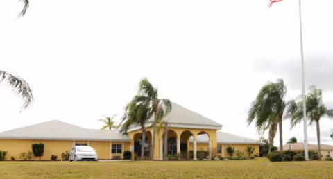 Single-story beige assisted living building with an arched entrance, palm trees, flagpole and a lawn.