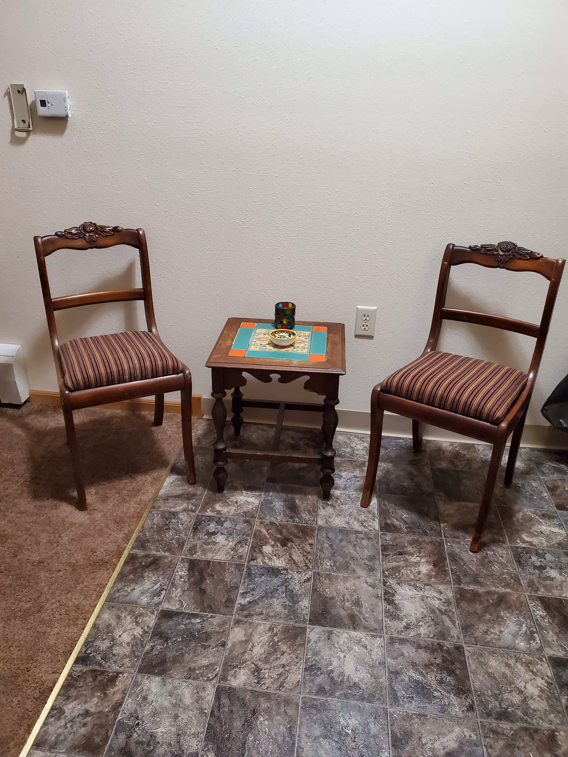 Two wooden chairs with striped cushions are placed on either side of a small wooden table with a colorful tiled top. The table holds a small decorative bowl and a multicolored glass. The floor is partially carpeted and partially covered with dark patterned tiles. The wall behind is plain with a power outlet and a thermostat or control panel.