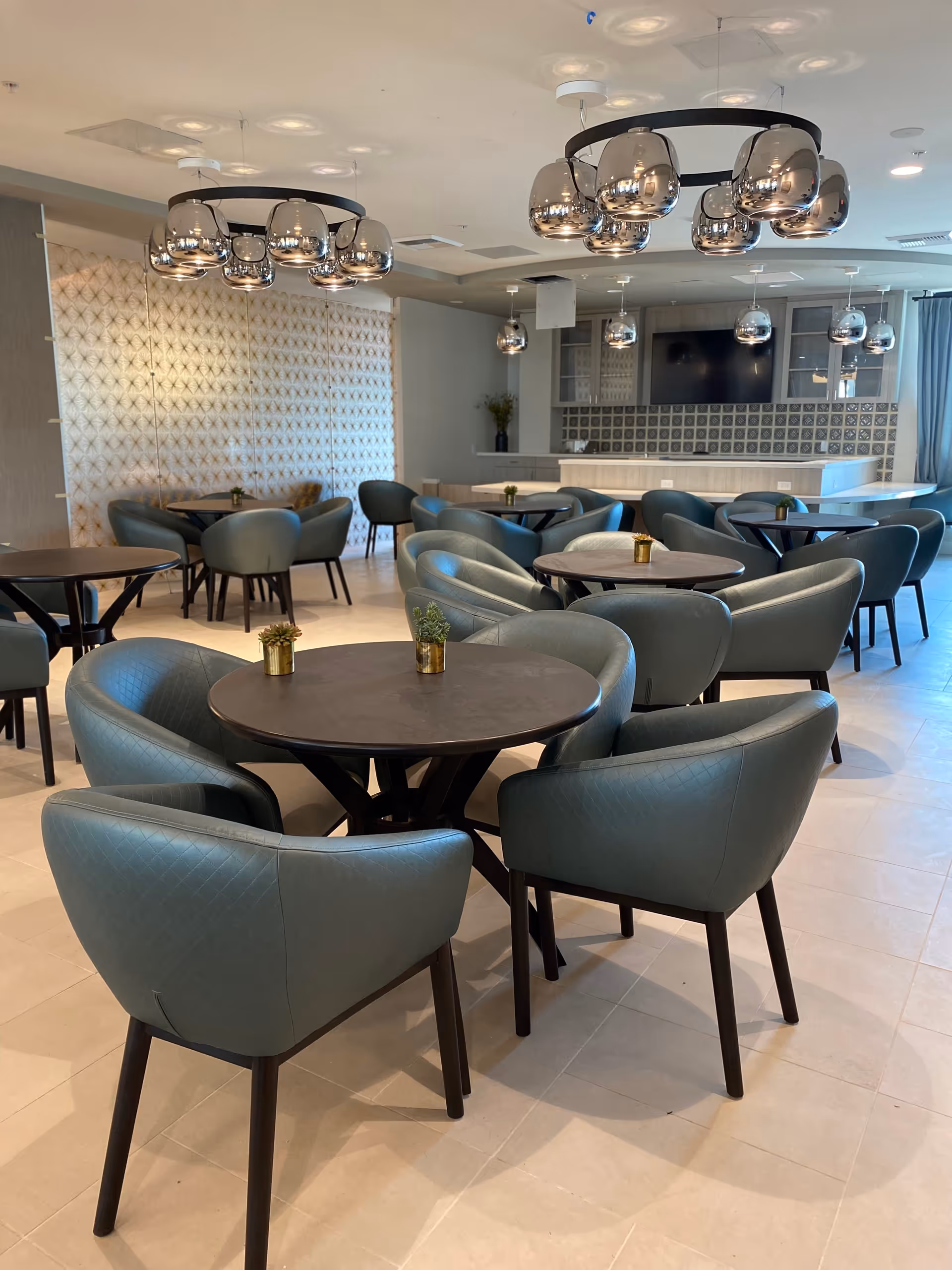 A modern dining area with multiple round tables surrounded by teal upholstered chairs. The room features contemporary pendant lights with reflective metallic finishes hanging from the ceiling. In the background, there is a counter area with a tiled backsplash and a mounted flat-screen TV. The space is well-lit with natural light coming from windows on the right side.