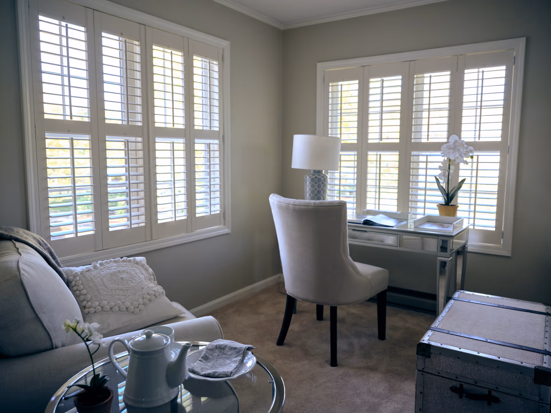 A cozy living room with large windows covered by white plantation shutters. The room features a light-colored sofa with a decorative pillow, a glass-top side table with a teapot and a small plant, a cushioned chair at a mirrored desk with a lamp and a potted orchid, and a vintage-style trunk on the carpeted floor.