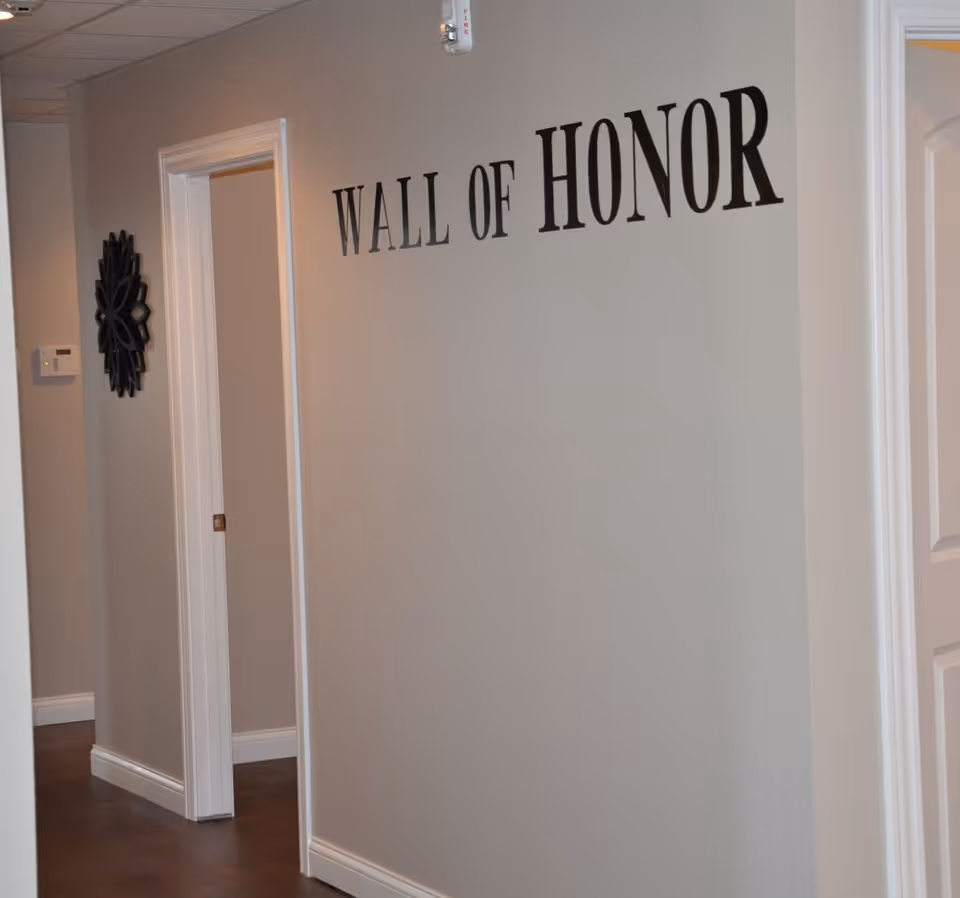 Interior hallway with beige walls and white trim featuring the words 'WALL OF HONOR' in large black letters on one wall. There are doorways on either side of the hallway and a decorative black wall piece near a thermostat.