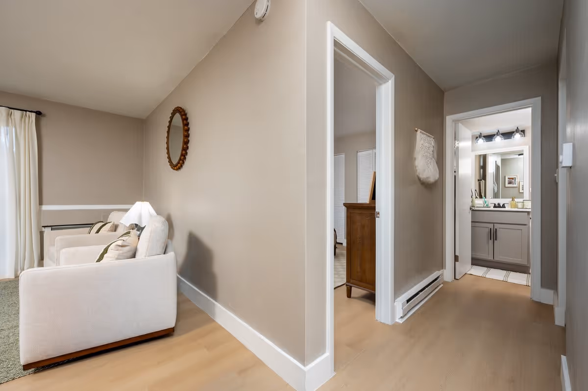 Interior view of a senior living facility showing a hallway with light wood flooring and beige walls. To the left, there are two beige armchairs with cushions and a lamp between them, next to a window with white curtains. A round decorative mirror hangs on the wall above the chairs. The hallway leads to two open doorways, one showing a room with a wooden dresser and the other revealing a bathroom with a vanity, mirror, and three light fixtures above it.