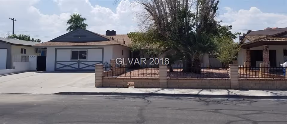 Front exterior of a single-story house with a driveway, low brick wall and desert landscaping under a partly cloudy sky.