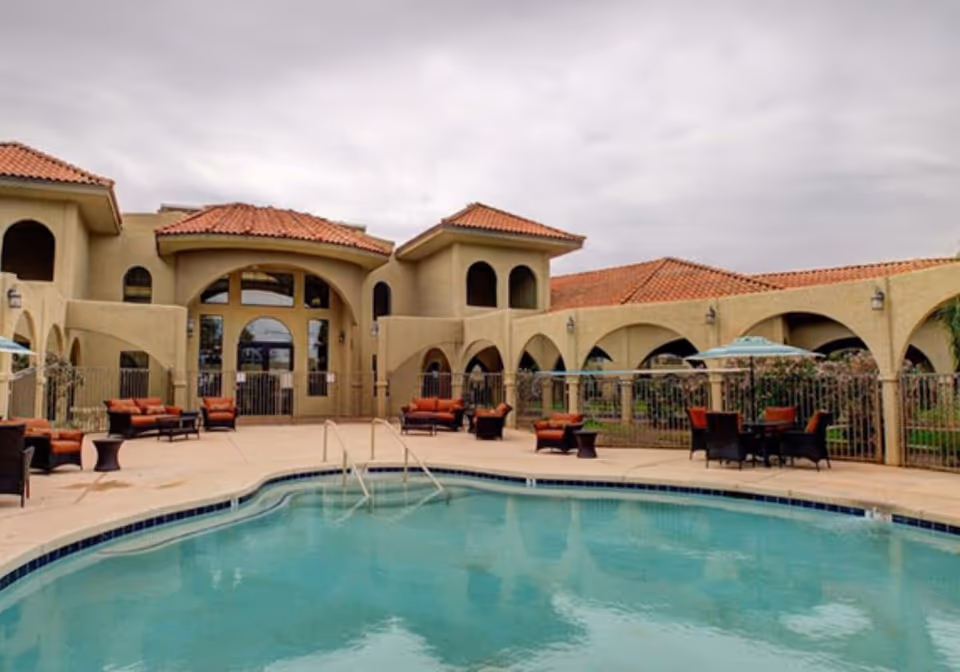 Outdoor swimming pool area with patio furniture including chairs and tables with umbrellas, surrounded by a beige building with arches and red tile roofs under a cloudy sky.