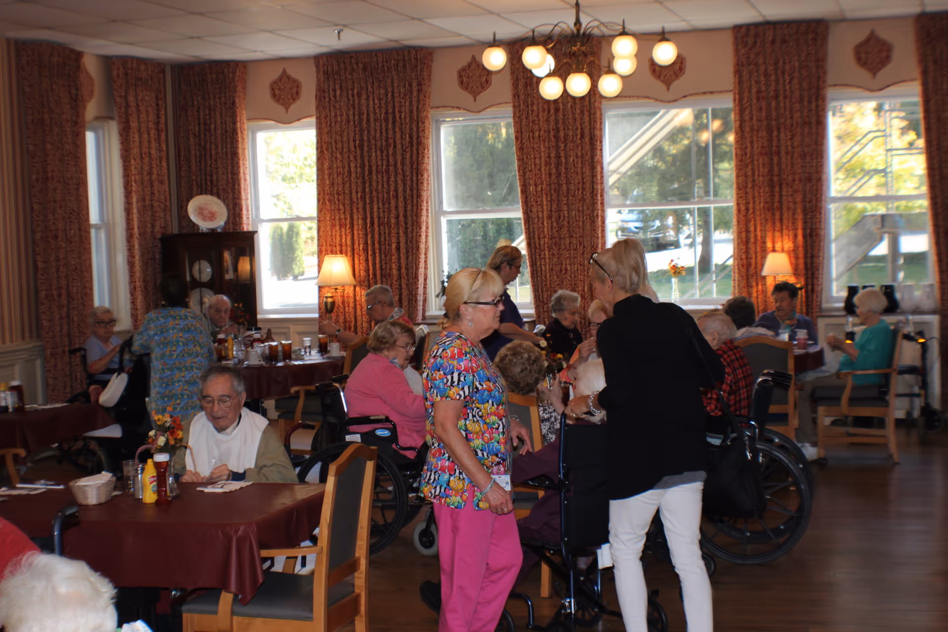 A dining room in an assisted living facility with several elderly residents seated at tables covered with maroon tablecloths. Some residents are in wheelchairs, and a few staff members are interacting with them. The room has large windows with patterned curtains, wooden floors, and warm lighting from lamps and a ceiling fixture.