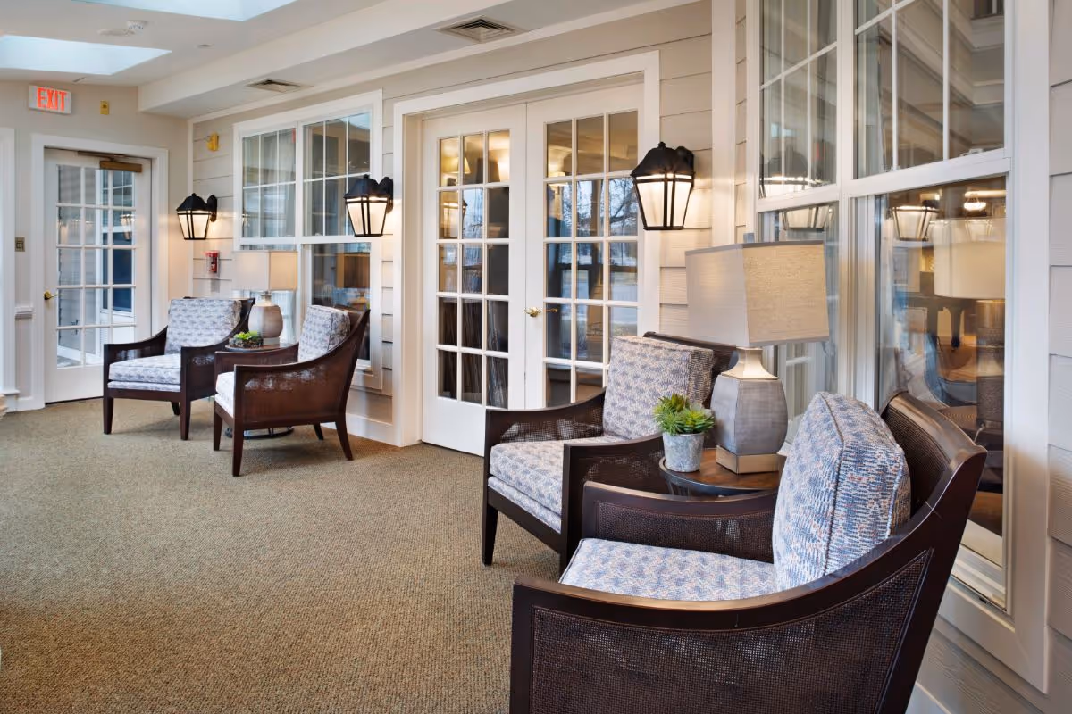 Sunlit common seating area with upholstered chairs, side tables and lamps along a wall of French doors and windows.