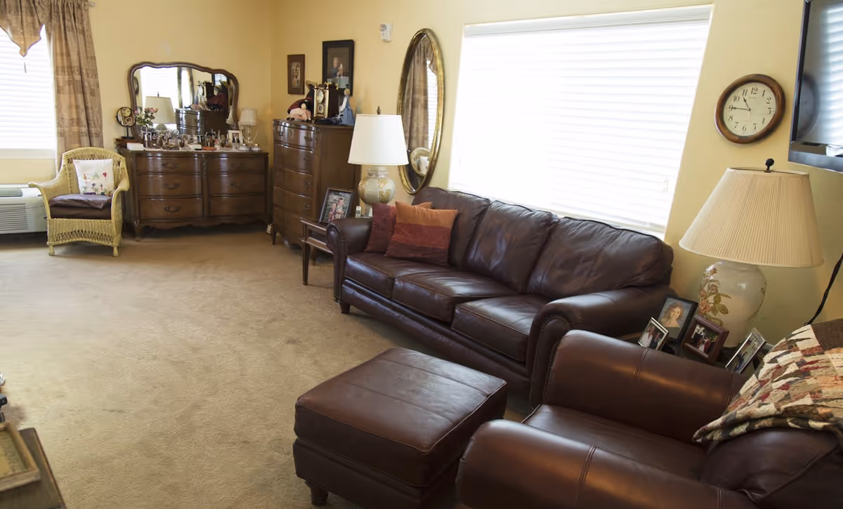 A cozy living room with a brown leather sofa, matching armchair, and ottoman. There is a large window with blinds, a round wall clock, a table lamp on a side table with framed photos, and a dresser with a mirror and various decorative items. A wicker chair with a cushion is placed near another window with curtains.