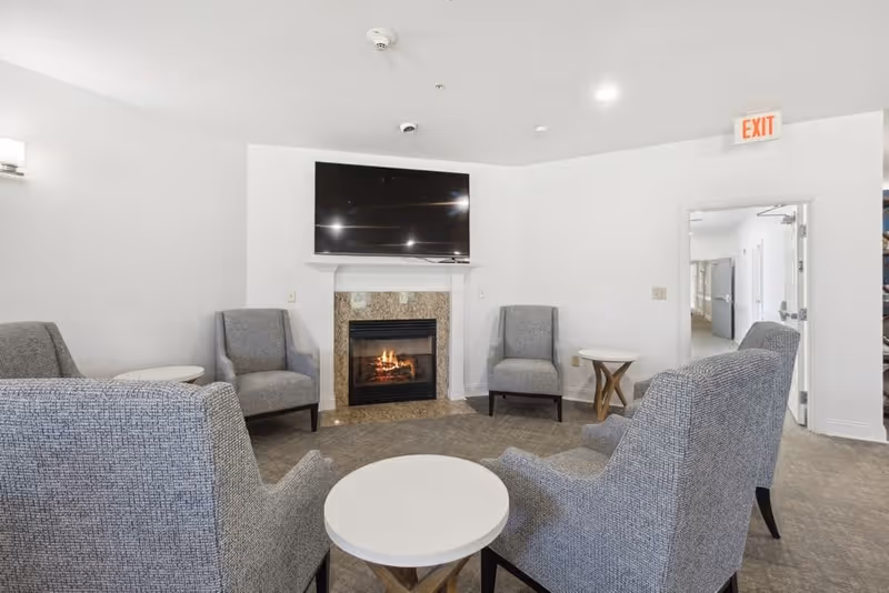 A cozy seating area with five gray upholstered armchairs arranged around two small round white tables. A fireplace with a granite surround is centered on the far wall, above which a large flat-screen TV is mounted. The walls are white, and there is an exit sign above an open door leading to a hallway.