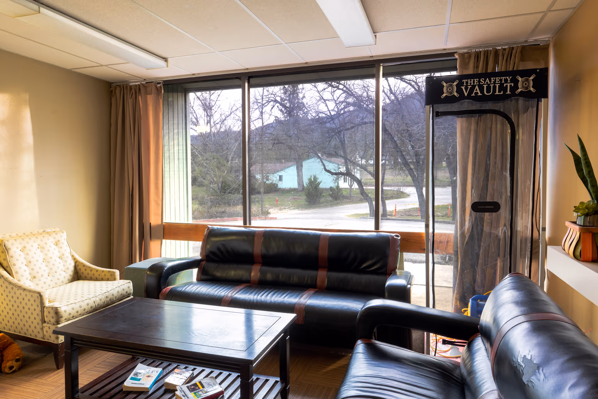 A sunlit living room with black leather sofas and an upholstered armchair around a coffee table in front of large windows overlooking trees.