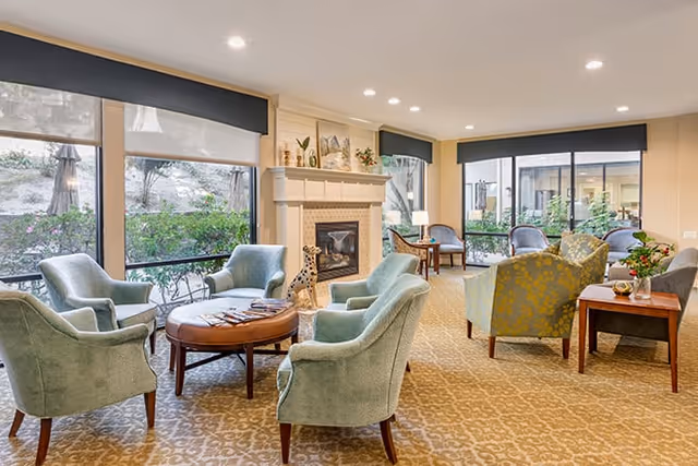 A cozy living room area with several upholstered armchairs arranged around a round wooden coffee table. There is a fireplace with decorative items on the mantel, large windows with black valances letting in natural light, and a patterned carpet covering the floor. Outside the windows, greenery and patio umbrellas are visible.