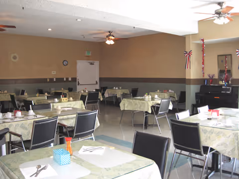 Interior view of a dining room with multiple tables covered with light green patterned tablecloths. Each table is set with cups, napkins, and utensils. Black chairs surround the tables. The room has beige walls with a dark brown stripe and ceiling fans with lights. There is a piano in the corner decorated with red, white, and blue ribbons.
