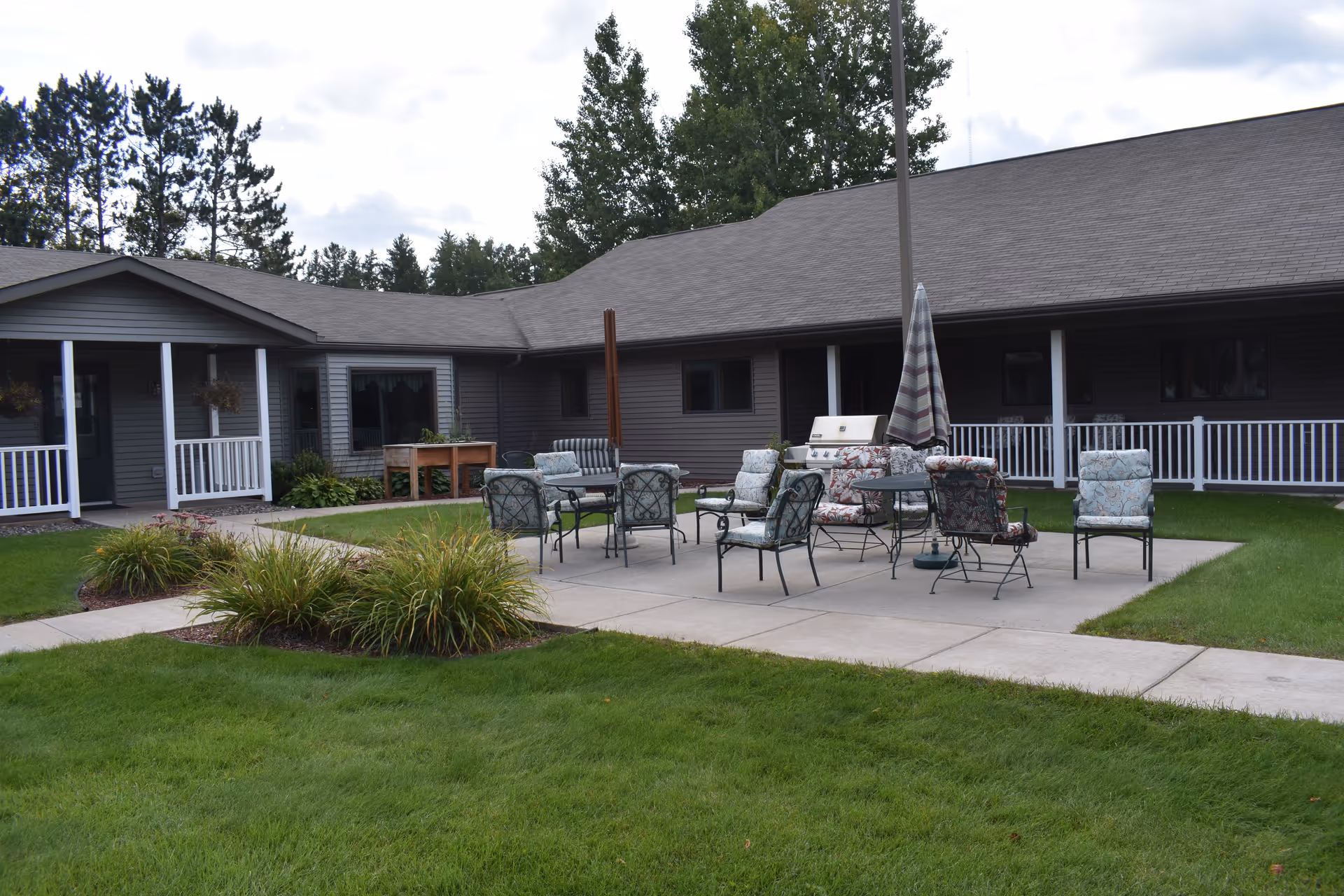 Outdoor patio area at Harmony House Brainerd with several cushioned chairs and tables arranged on a concrete slab surrounded by green grass and landscaping. The building with a covered porch and white railing is visible in the background under a cloudy sky.