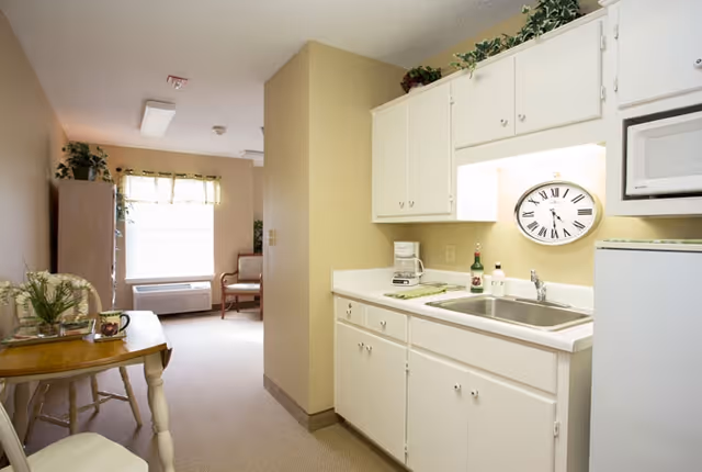 A small kitchen area with white cabinets, a sink, a coffee maker, and a microwave. There is a large clock on the wall above the sink. To the left, a small dining table with chairs is visible, and further back, a window with a valance and a chair near an air conditioning unit can be seen.