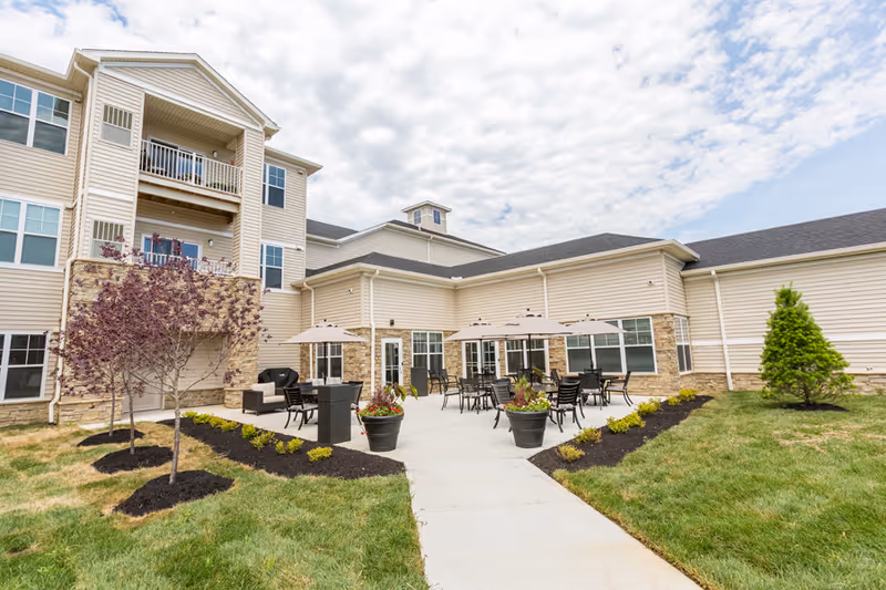 Outdoor patio area of a senior living facility with tables, chairs, and umbrellas on a concrete surface. The building has beige siding with stone accents and multiple windows. There are small landscaped areas with grass, shrubs, and young trees surrounding the patio. The sky is partly cloudy.
