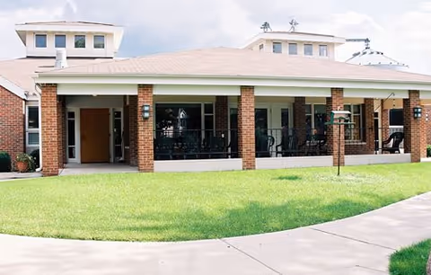 Exterior front of a brick building with a covered porch and patio chairs facing a green lawn and sidewalk.