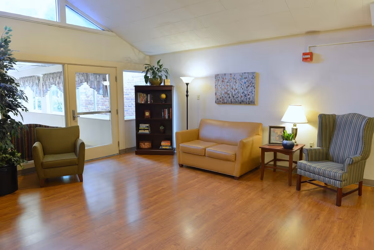A cozy living room area with a tan leather loveseat, a green armchair, and a blue-striped wingback chair. There is a wooden side table with a lamp, a framed photo, and a small plant. A tall floor lamp stands next to a bookshelf filled with books and decorative items. The room has wood flooring, a large window, and a glass door leading to a sunroom or enclosed porch.
