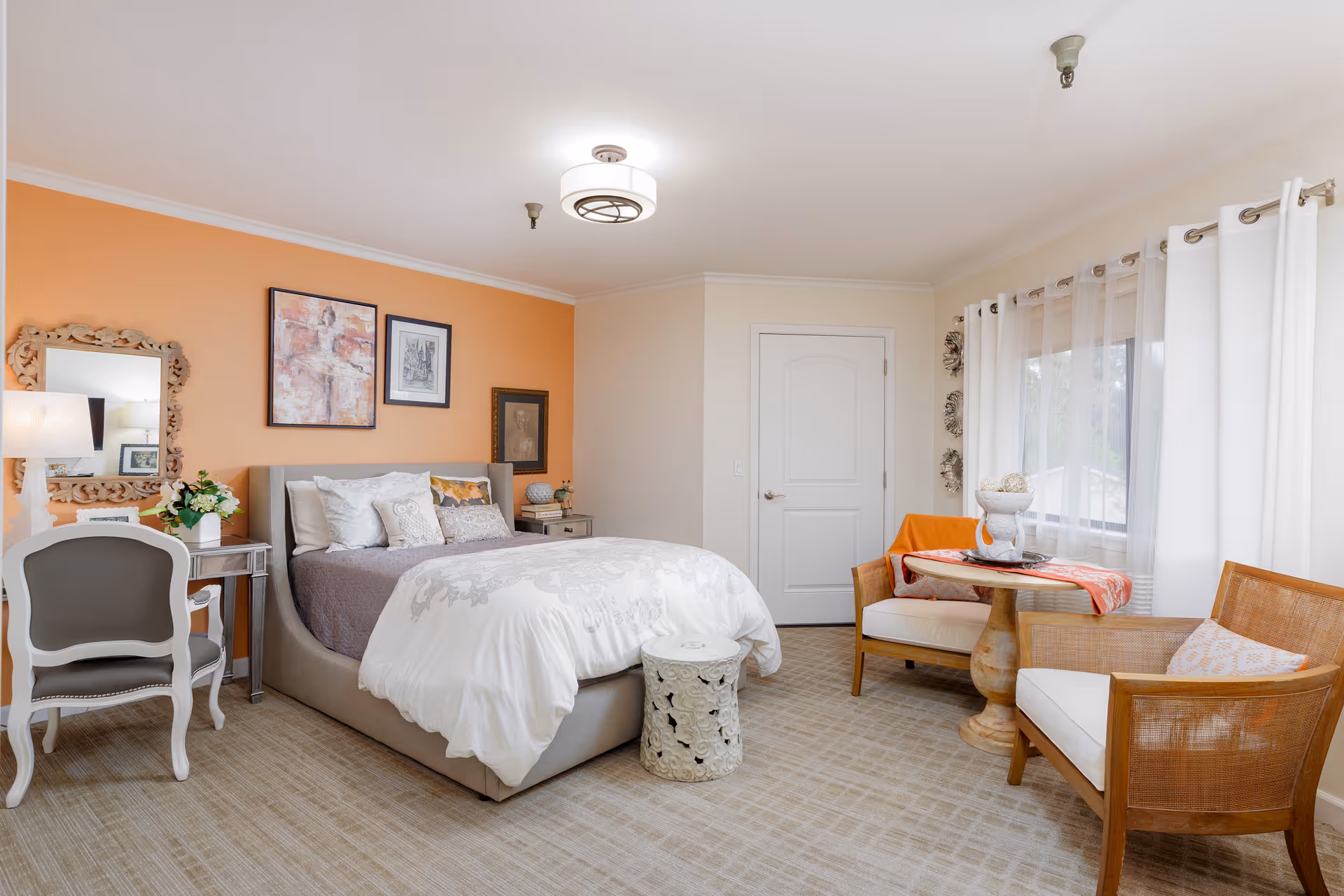 A bright and cozy bedroom with a bed featuring white and gray bedding, an orange accent wall with framed artwork, a small white desk with a chair and lamp, a round side table, and a sitting area with two wooden chairs and a small round table near a window with white curtains.