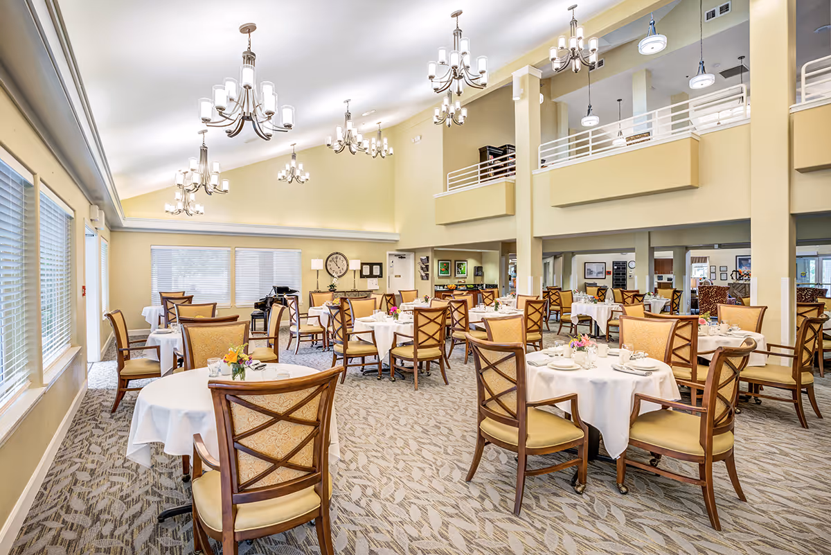 Bright dining room with round tables set with white linens and wooden chairs beneath multiple chandeliers.