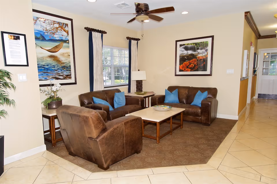 A cozy living room area with two brown leather sofas and one brown leather armchair arranged around a rectangular coffee table. The sofas have blue throw pillows. The walls are beige with two framed pictures, one of a hammock over water and another of a waterfall with orange flowers. There is a window with curtains and a ceiling fan above. The floor is tiled with a brown carpet under the seating area.