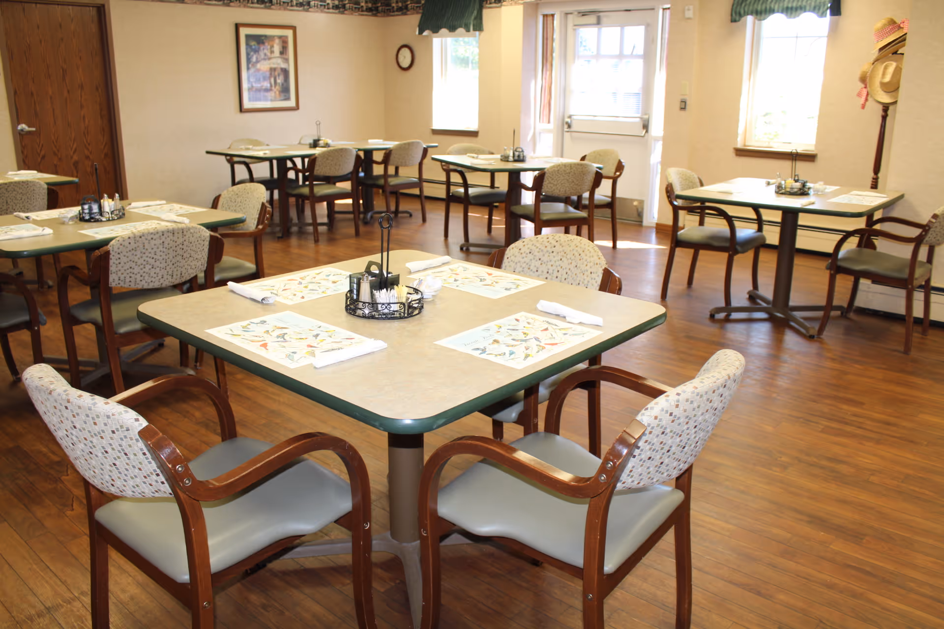 A dining room in a senior living facility with multiple square tables, each set with placemats, napkins, and condiment holders. The room has wooden flooring, beige walls, and several windows letting in natural light. Chairs with wooden arms and patterned upholstery surround the tables. A coat rack with hats is visible near a window.