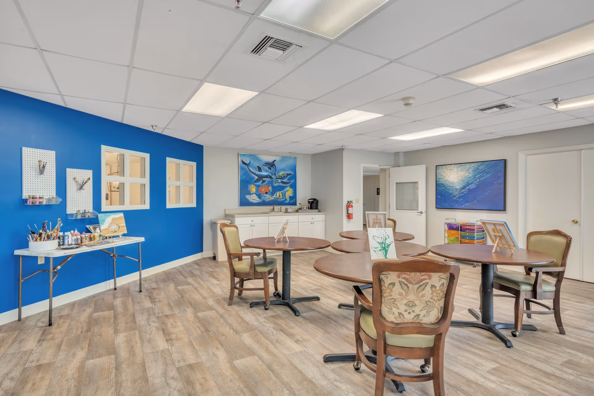 Bright activity room with round tables and chairs, art supplies on a table, and colorful paintings on the walls.