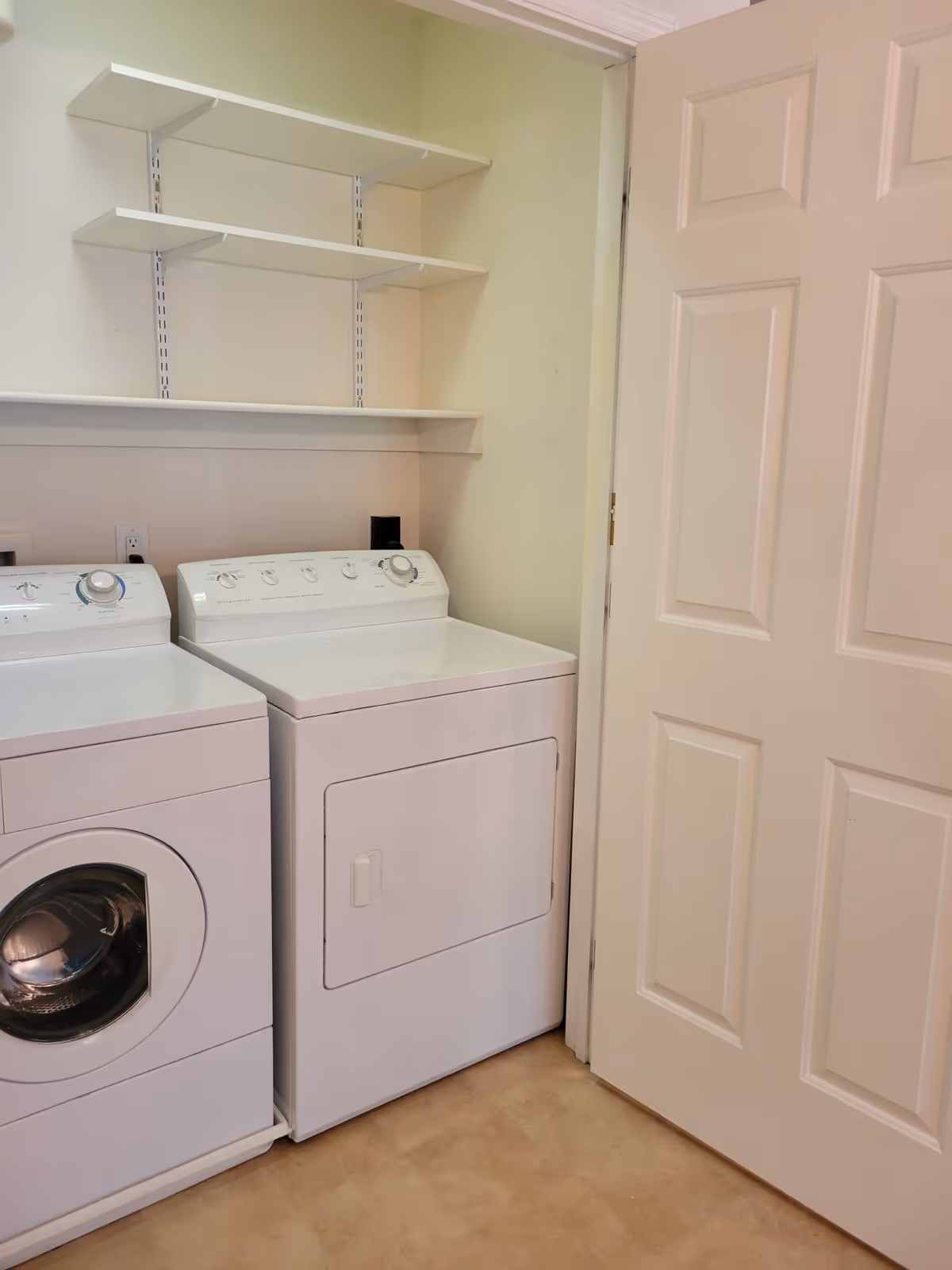Laundry room with a white front-loading washing machine and a white dryer side by side. Above the machines are three white wall-mounted shelves. A white door is partially open to the right.