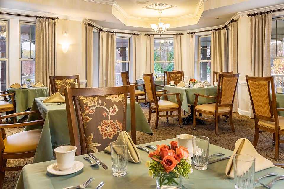 A bright dining room with multiple tables covered in green tablecloths, each set with beige napkins, glassware, and silverware. The chairs have wooden frames with floral-patterned upholstery. The room has large windows with beige curtains, allowing natural light to fill the space. A small floral centerpiece is on the nearest table.