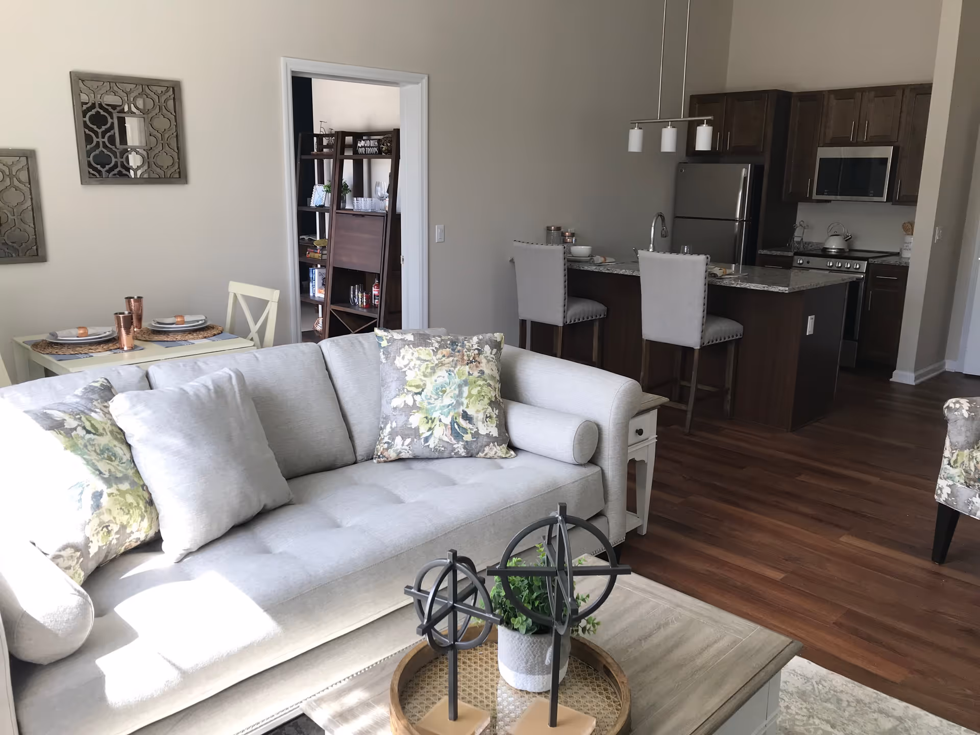 Open-plan living area featuring a light gray sofa with floral pillows, a coffee table, a small dining table, and a kitchen island with bar stools in the background.