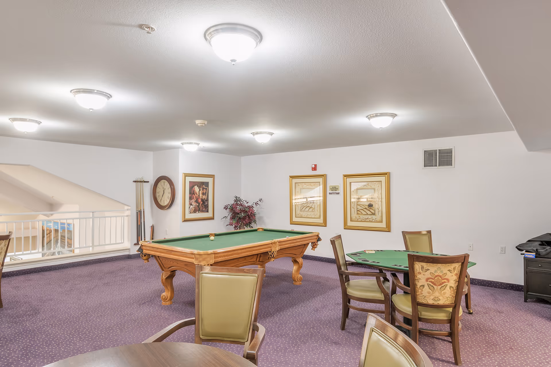A recreational room with a pool table and a card table surrounded by chairs. The room has purple carpet, white walls, framed artwork, a large wall clock, and ceiling lights. There is a railing overlooking a lower level on the left side.