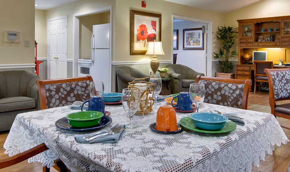 A dining table set with colorful dishes and glassware in a cozy senior living common area with sofas and a desk in the background.