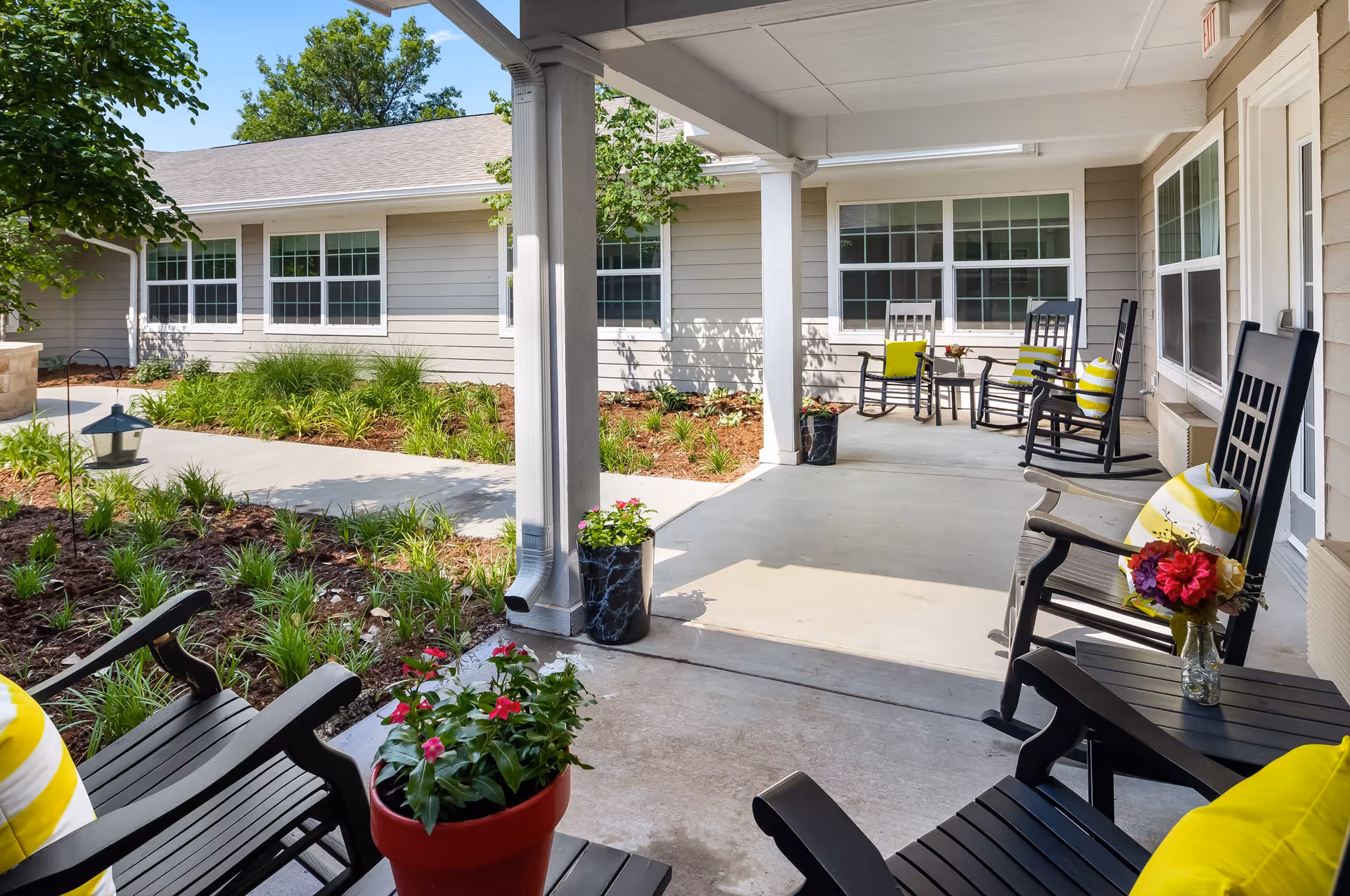 Covered outdoor patio area with black rocking chairs adorned with yellow and white striped cushions and small tables with flower vases. The patio overlooks a landscaped garden with green plants and a concrete walkway. The building exterior is light gray with white trim and multiple windows.