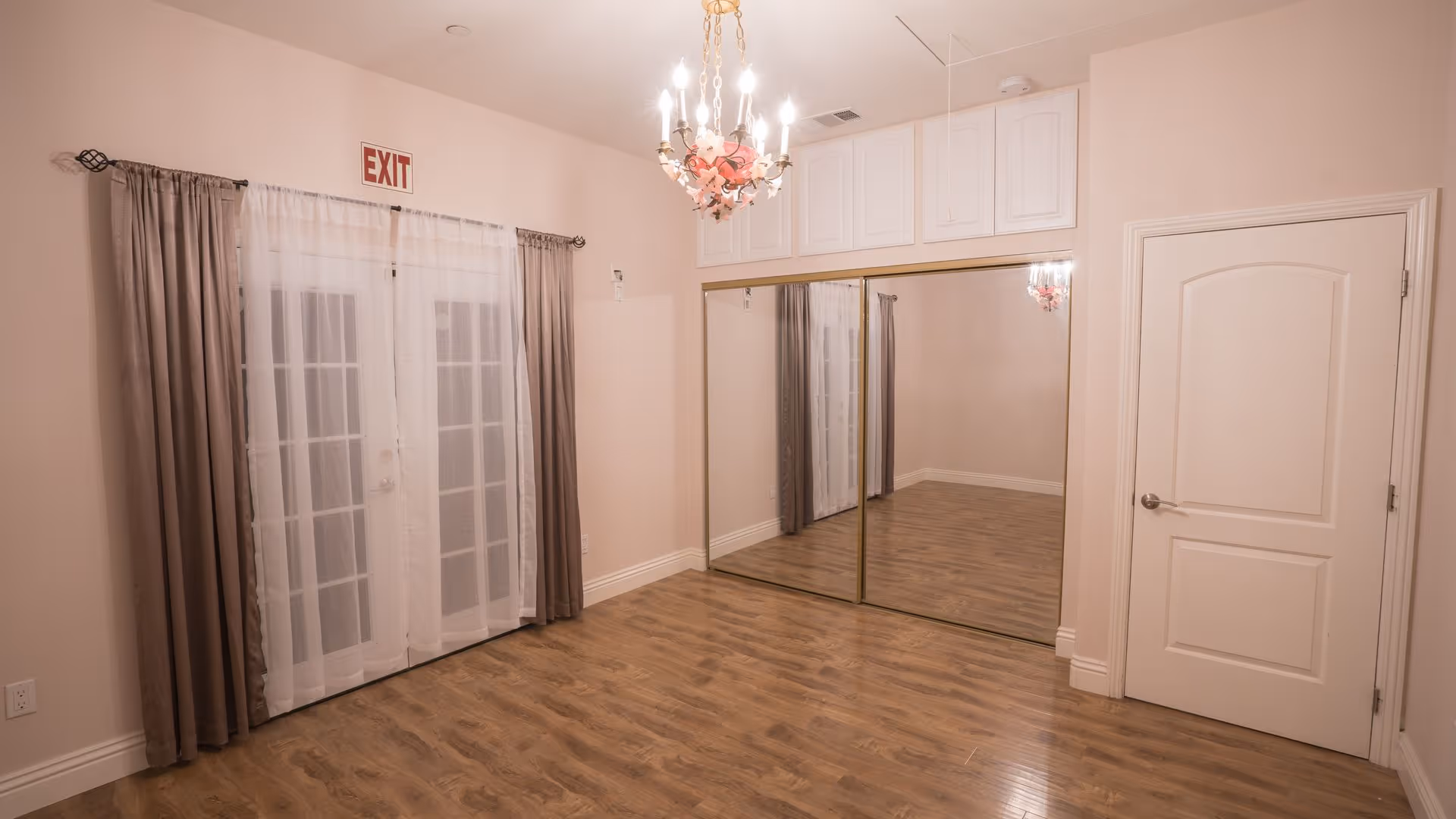 Empty room with wooden flooring, a chandelier hanging from the ceiling, a set of double glass doors with curtains and an exit sign above, a large mirrored closet with sliding doors, and a closed white door on the right wall.