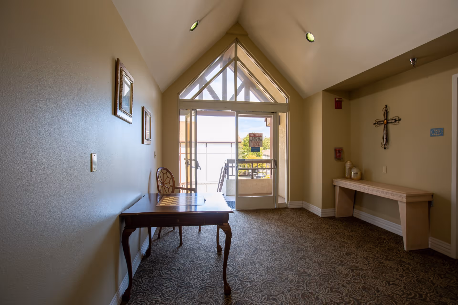 A small seating area in a hallway with a wooden table and chair on the left, a decorative cross on the right wall above a narrow console table, and a glass door leading outside under a triangular window letting in natural light.