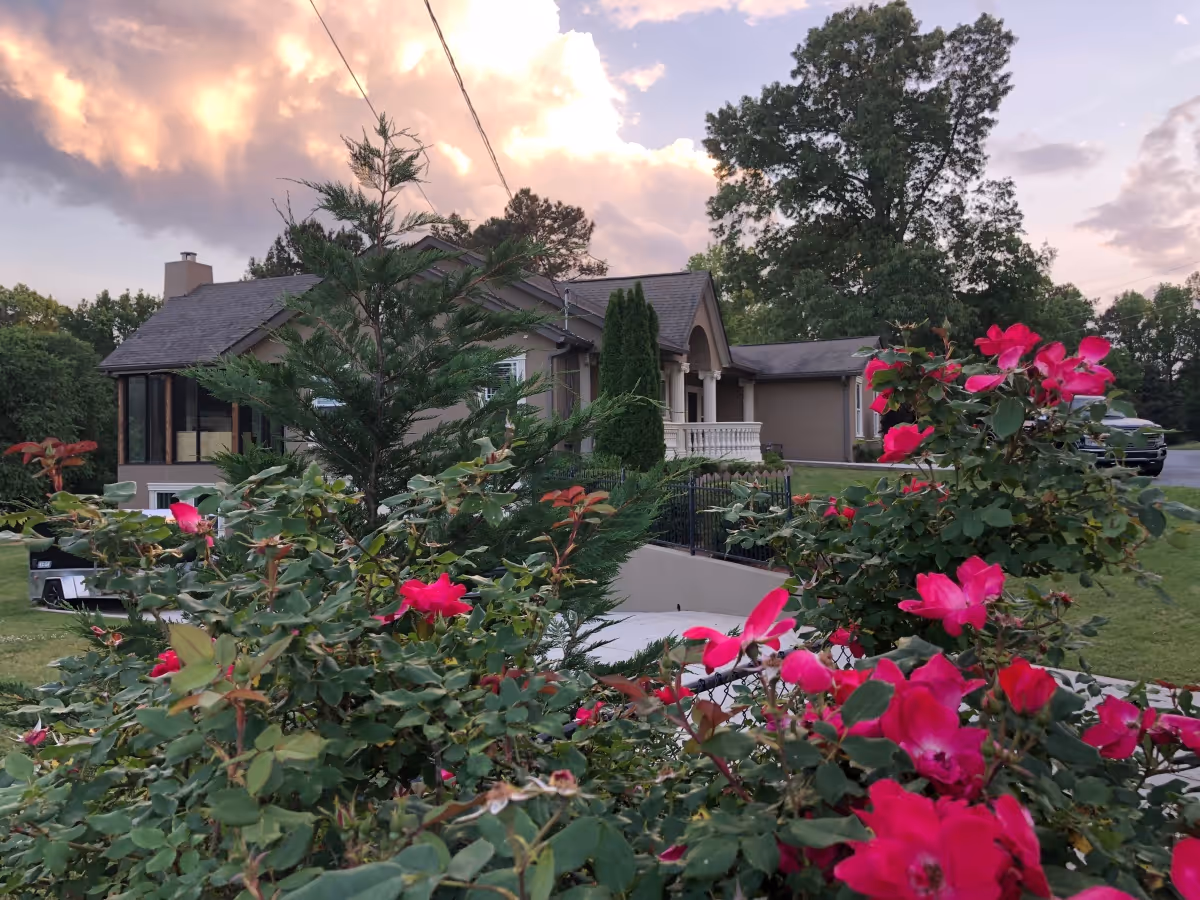 Exterior view of a senior care facility building surrounded by greenery and vibrant pink flowers in the foreground, with a partly cloudy sky at sunset.