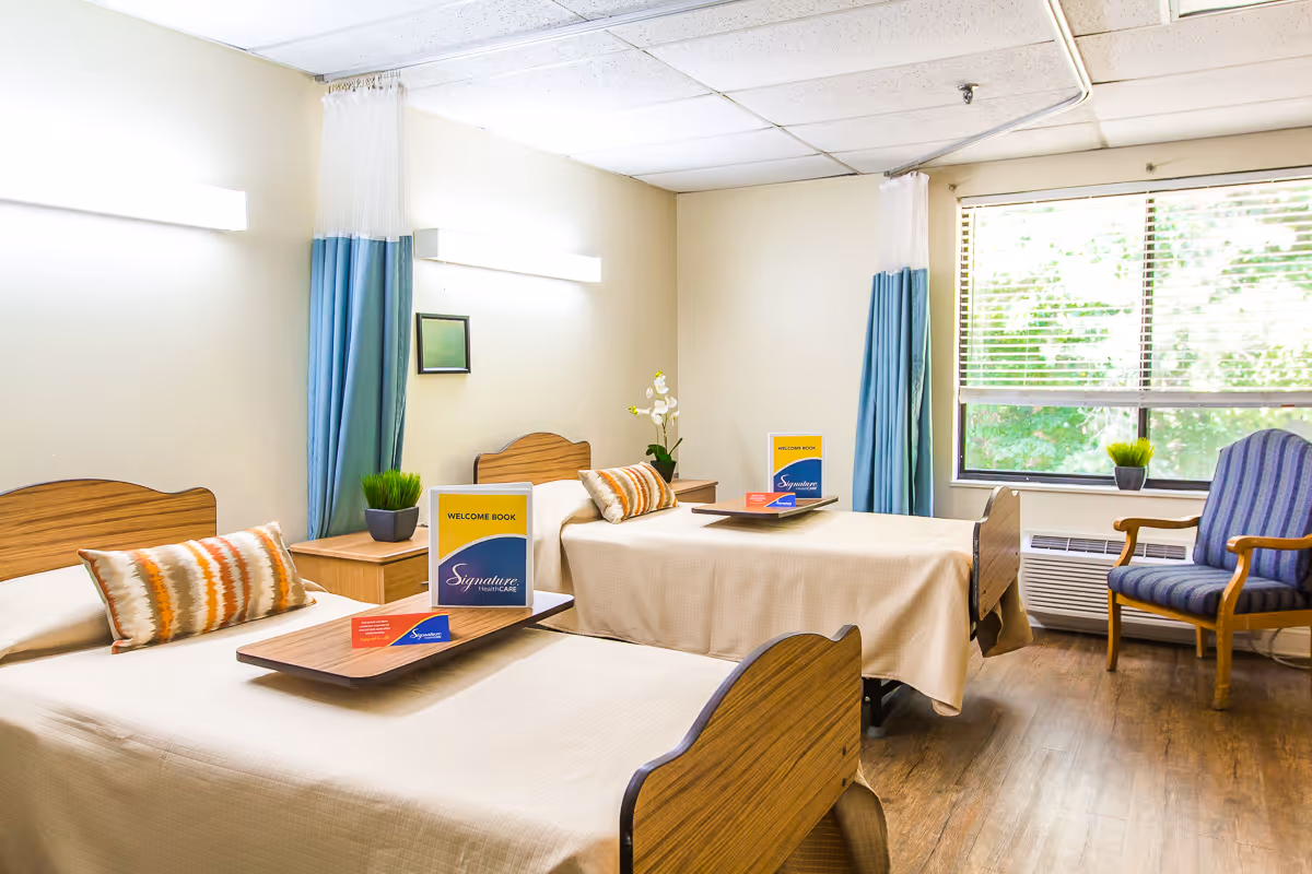 A bright and clean room with two single beds, each with a beige bedspread and a decorative pillow. Each bed has a wooden tray with a welcome book and pamphlet. There is a small wooden nightstand with a potted plant between the beds. Blue and white privacy curtains hang on the walls behind the beds. A large window with blinds lets in natural light, and a blue upholstered chair with wooden arms is positioned near the window. The floor is wood, and the walls are painted a light cream color.