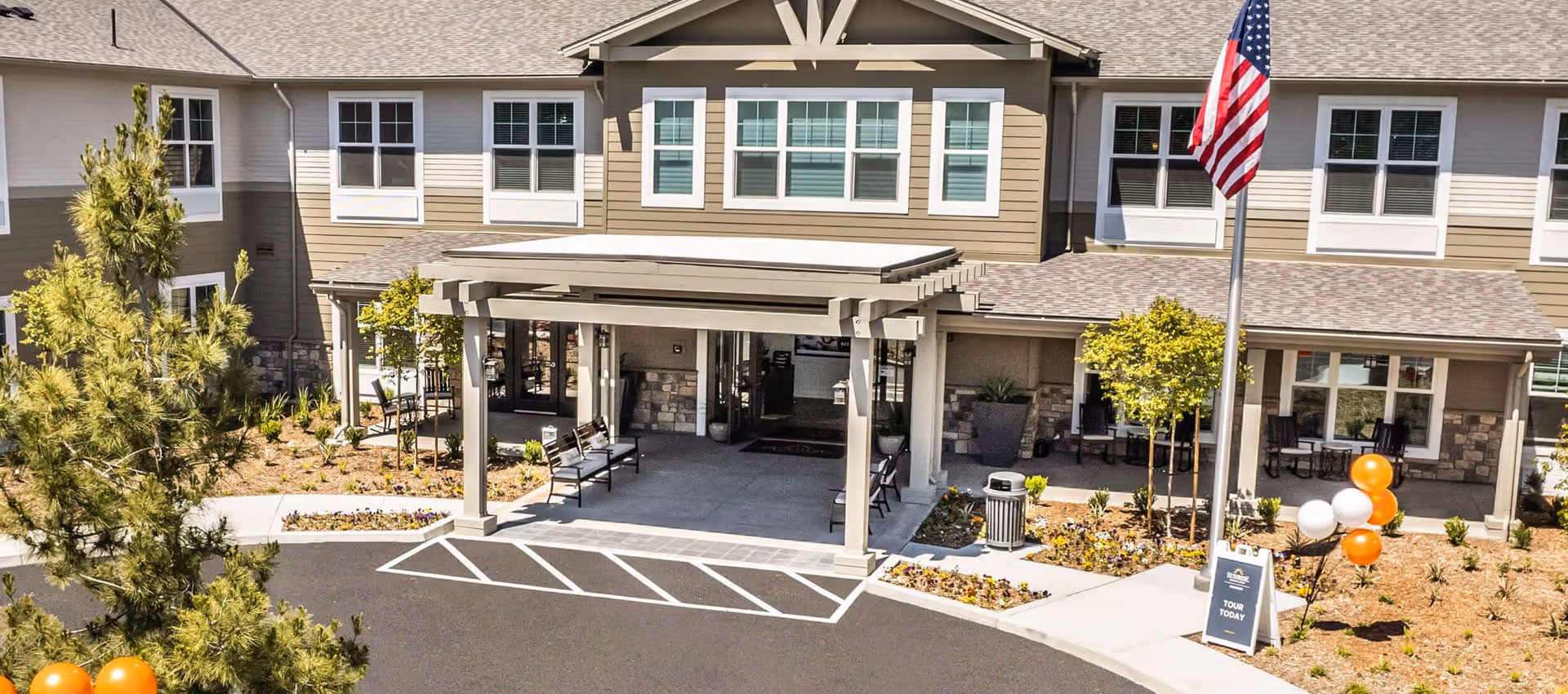 Front exterior view of a senior living facility named Sunrise of Oceanside with a covered entrance, benches, landscaped garden beds, an American flag, and balloons near a sign that says 'Tour Today'.