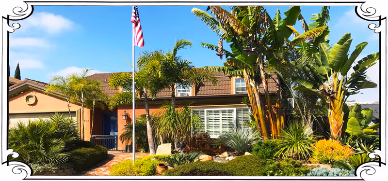 Front exterior view of a single-story house with a brown tiled roof, surrounded by lush tropical plants and palm trees. An American flag is flying on a flagpole in the front yard under a clear blue sky.