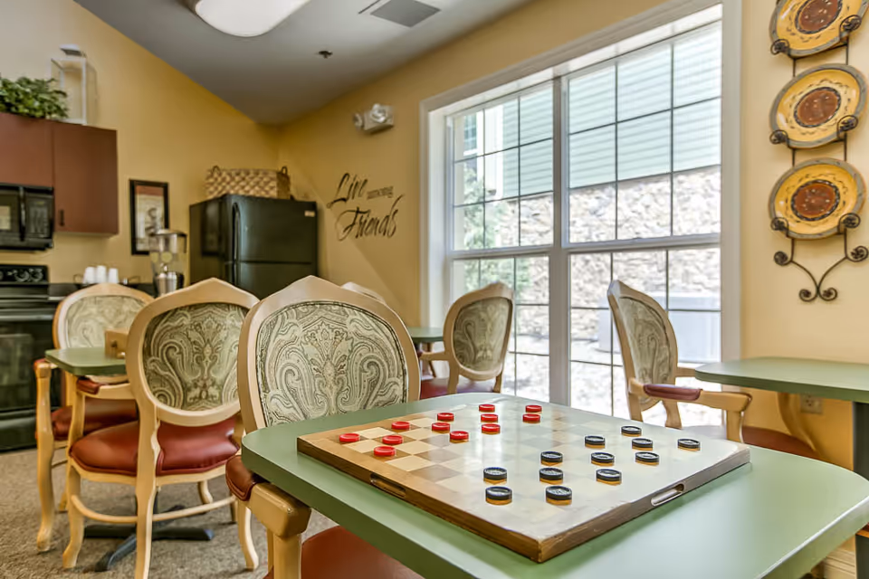 A cozy room with several tables and upholstered chairs, featuring a checkers game set up on the nearest table. The room has a large window letting in natural light, a black refrigerator, a microwave, and decorative plates on the wall. The wall also has a decal that reads 'Live with Friends.'