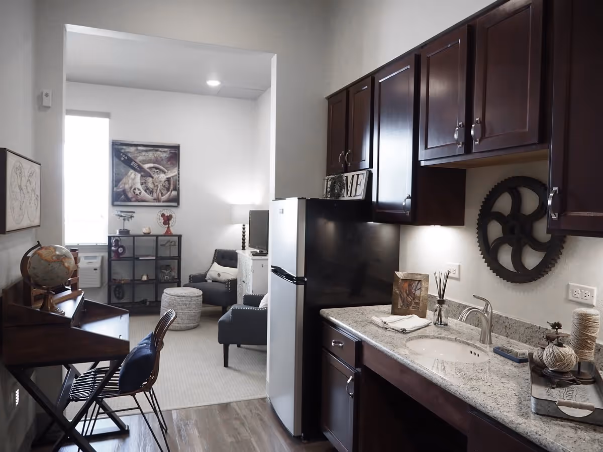 Interior view of a senior living facility unit showing a small kitchenette with dark wood cabinets, a granite countertop with a sink, and decorative items. Adjacent to the kitchenette is a living area with a gray armchair, a small ottoman, a TV on a stand, and a shelving unit with various decorations. A wooden desk with a globe and a chair is also visible. The room has light-colored walls and flooring, with a window providing natural light.