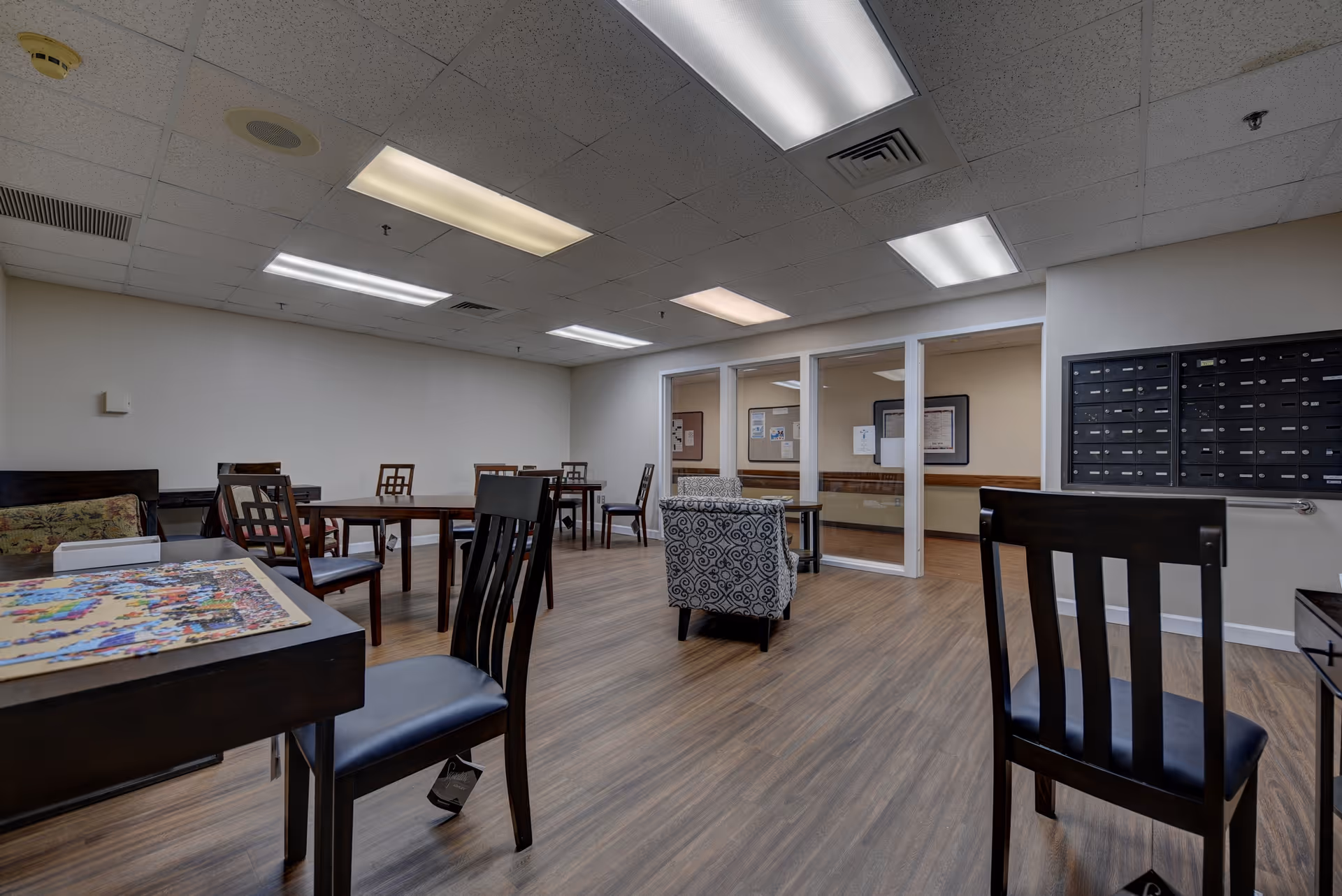 A spacious room with wooden flooring and multiple tables and chairs arranged for seating. There is a patterned armchair in the center near glass windows that look into another room. On the right wall, there is a set of mailboxes. The ceiling has several fluorescent lights providing bright illumination.