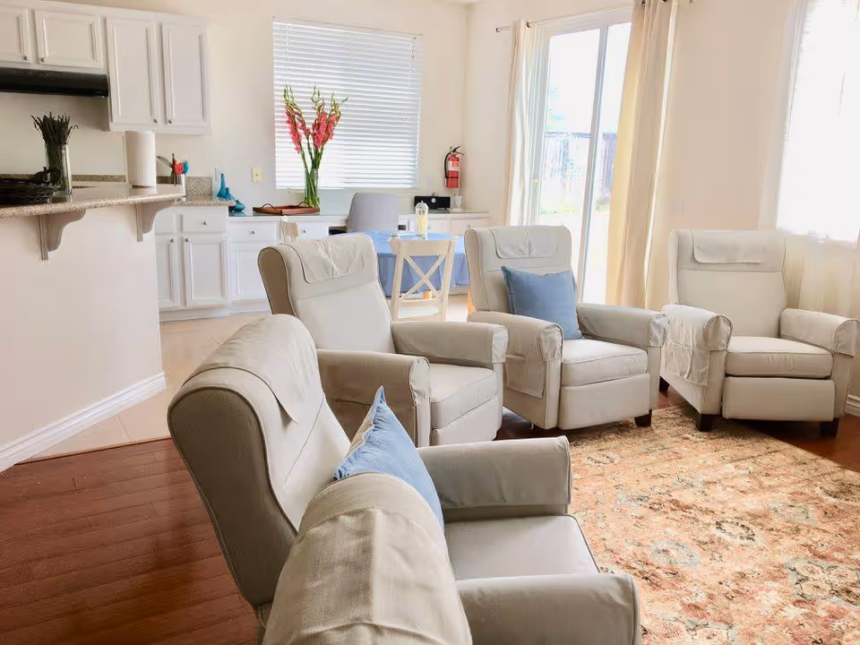 Bright communal living room with four beige recliner chairs around a patterned rug, a small table and chairs, and a kitchenette in the background.