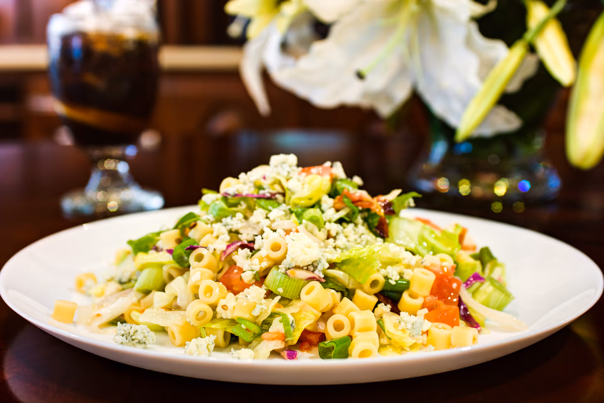 Close-up of a plate of pasta salad with diced vegetables and crumbled cheese on a wooden table, with a glass of iced drink and white flowers in the background.