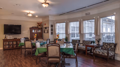 A well-lit common area with several tables covered in green tablecloths and surrounded by chairs. There are large windows with beige curtains letting in natural light. The room features a wooden floor, a flat-screen TV mounted on the wall, a wooden cabinet with decorative items, and a few armchairs arranged near the windows.