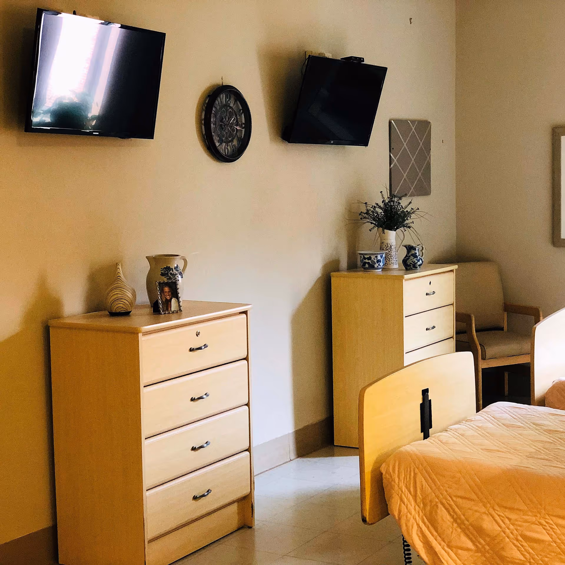 Interior view of a senior living facility room with two wooden dressers, each decorated with vases and a framed photo. Two wall-mounted flat-screen TVs and a decorative wall clock are visible above the dressers. A beige armchair is placed in the corner next to one dresser, and part of a bed with a peach-colored quilt is visible in the foreground.