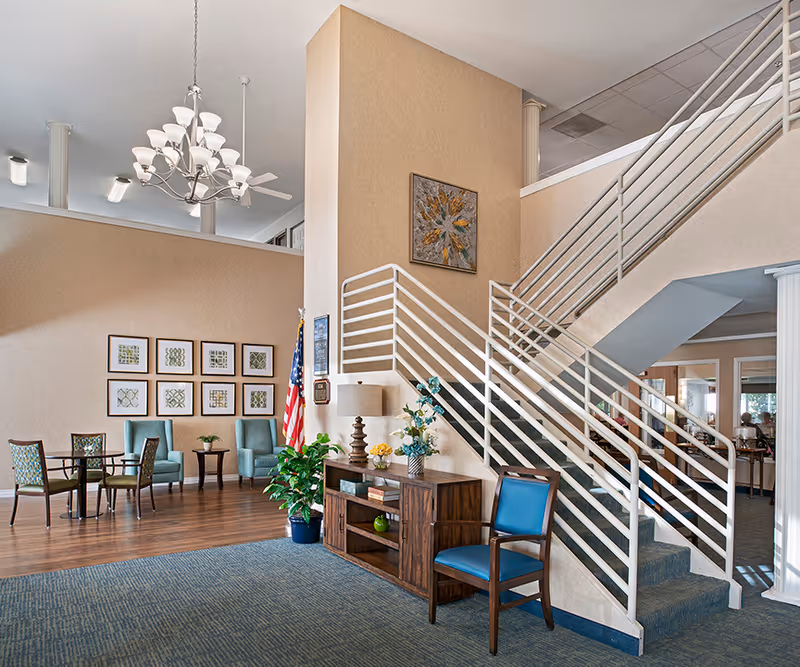 Interior view of a senior living facility lobby area with a staircase featuring white railings. There is a wooden console table with a lamp, flowers, and decorative items. A blue cushioned chair is placed next to the table. In the background, there are several framed artworks on the wall, an American flag, and a seating area with blue and patterned chairs around small tables. The ceiling has a chandelier and ceiling fans.