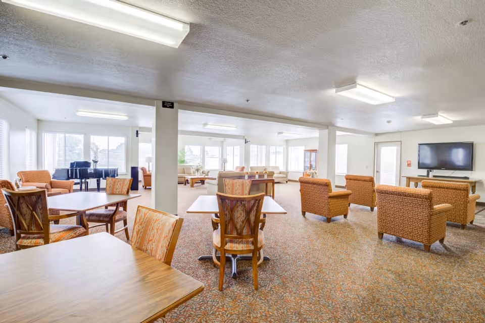 A spacious senior living common area with multiple seating arrangements including patterned armchairs and wooden tables with chairs. Large windows allow natural light to fill the room. A black grand piano is visible in the background near the windows. A flat-screen TV is mounted on the wall to the right. The room has a carpeted floor and fluorescent ceiling lights.