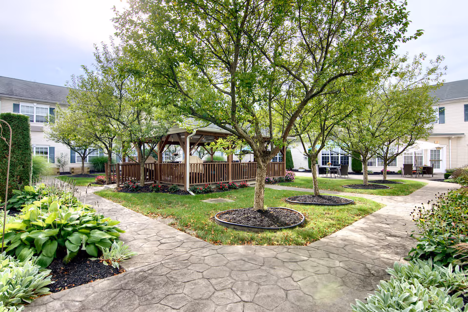 A landscaped courtyard with a wooden gazebo, several trees, paved walkways, and the surrounding senior living building.
